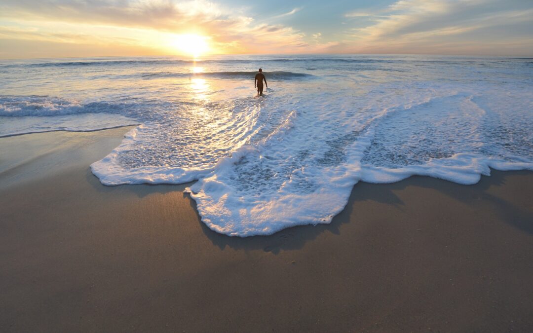 White foaming waves rolls on a sandy beach as the sun sets with a man walking in the water in the distance